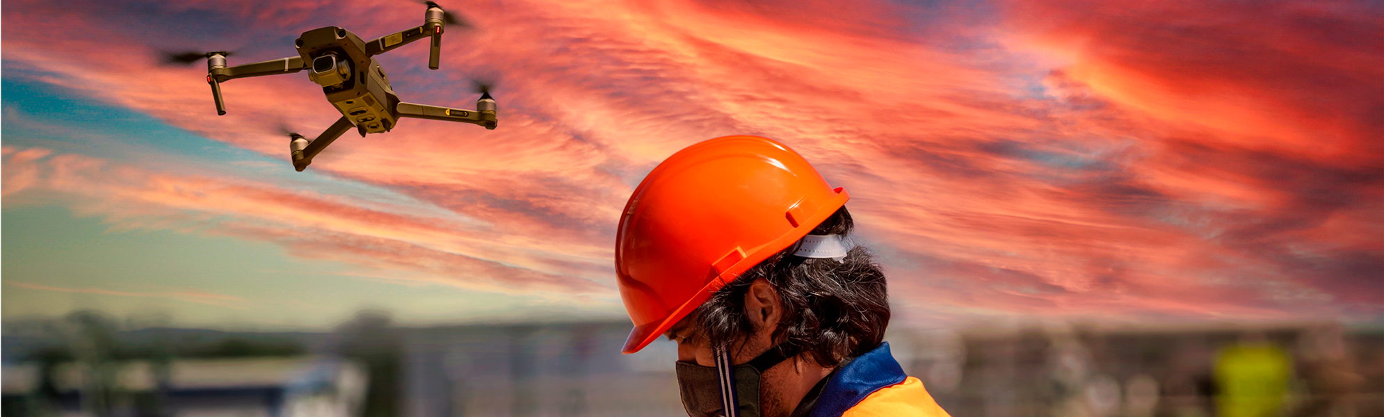 Ilunabarrean hegan egiten ari den drone baten irudia, segurtasun-kaskoa duen pertsona batek urrutiko kontrol bat erabiltzen duen bitartean
